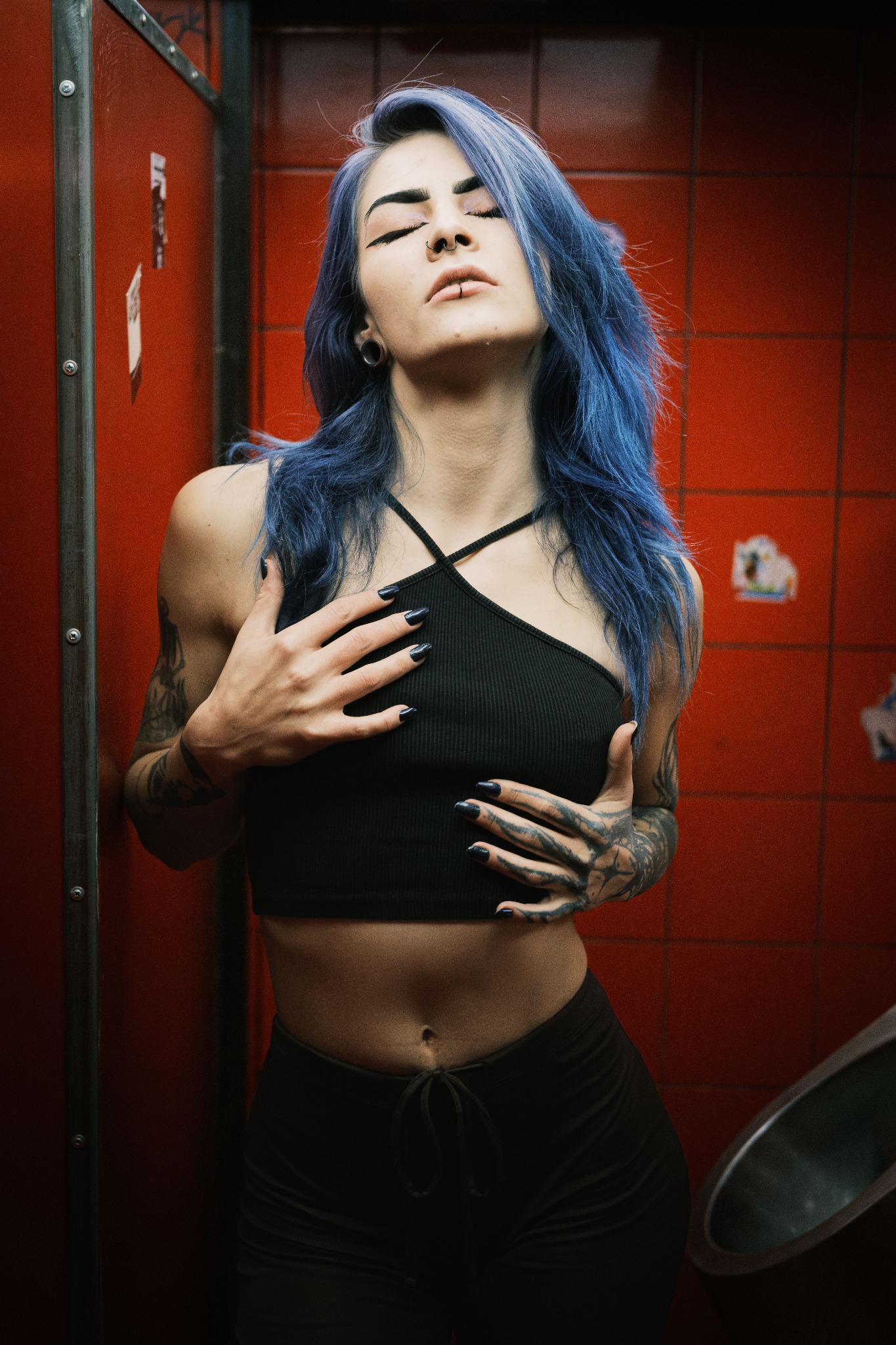 Kobieta z niebieskimi włosami i tatuażami pozuje w czerwonej łazience, z rękami na klatce piersiowej i zamkniętymi oczami.