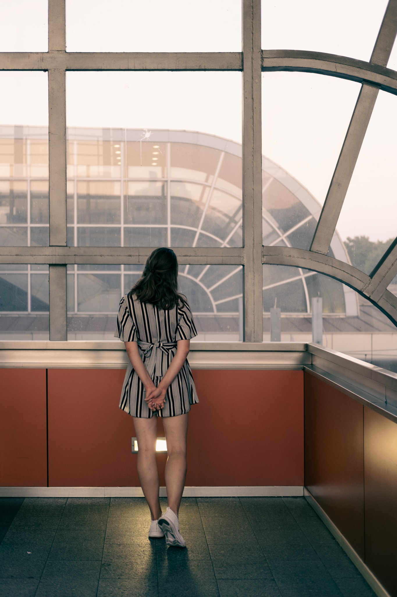 Kobieta w paski stoi tyłem, splecione dłonie, jasne buty sportowe, duże okno z widokiem na przeszklony budynek o łukowatej konstrukcji.