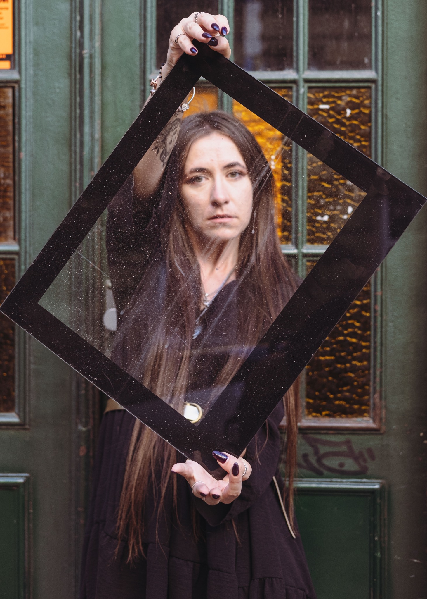 Kobieta z długimi włosami trzymająca czarną ramkę na tle zielonych drzwi z szybami