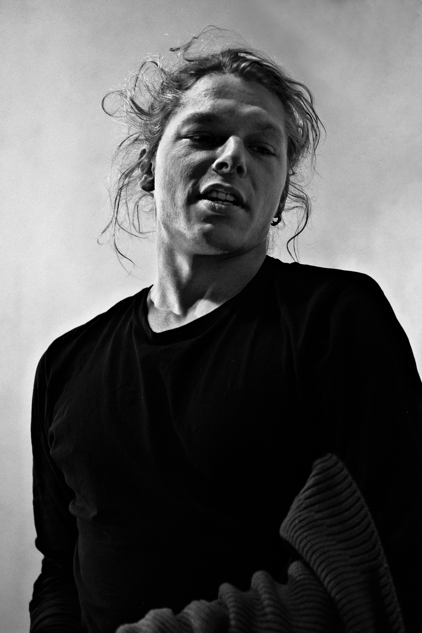 Czarno-białe zdjęcie portretowe mężczyzny z rozwianymi włosami, ujętego z dołu, z widoczną częścią czarnego swetra