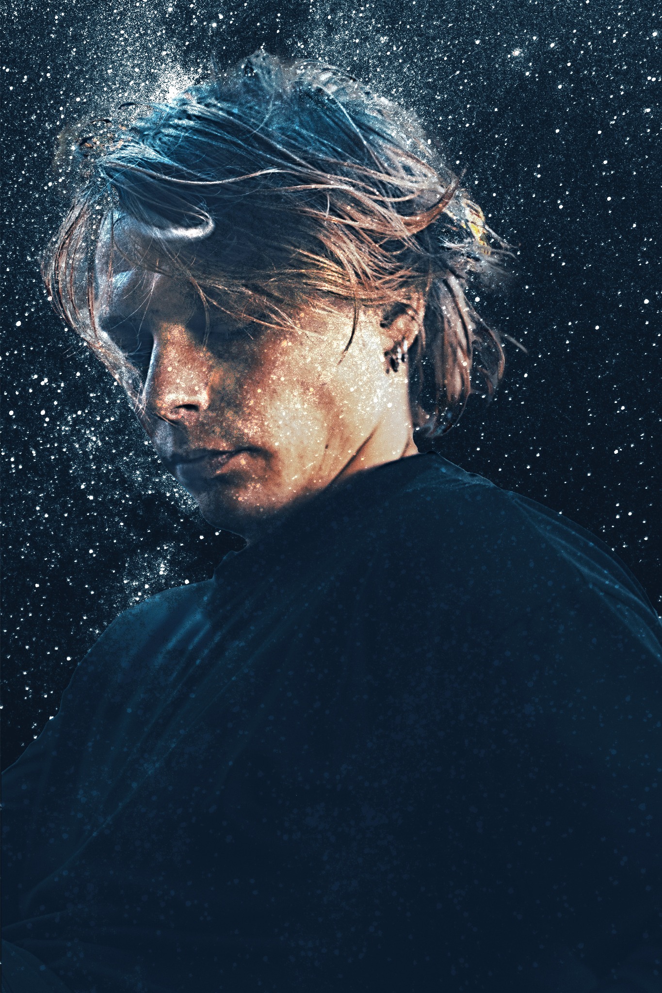 Portret młodego mężczyzny z kolczykiem, oświetlonego efektem przypominającym kosmiczny pył, wpatrującego się w dół.