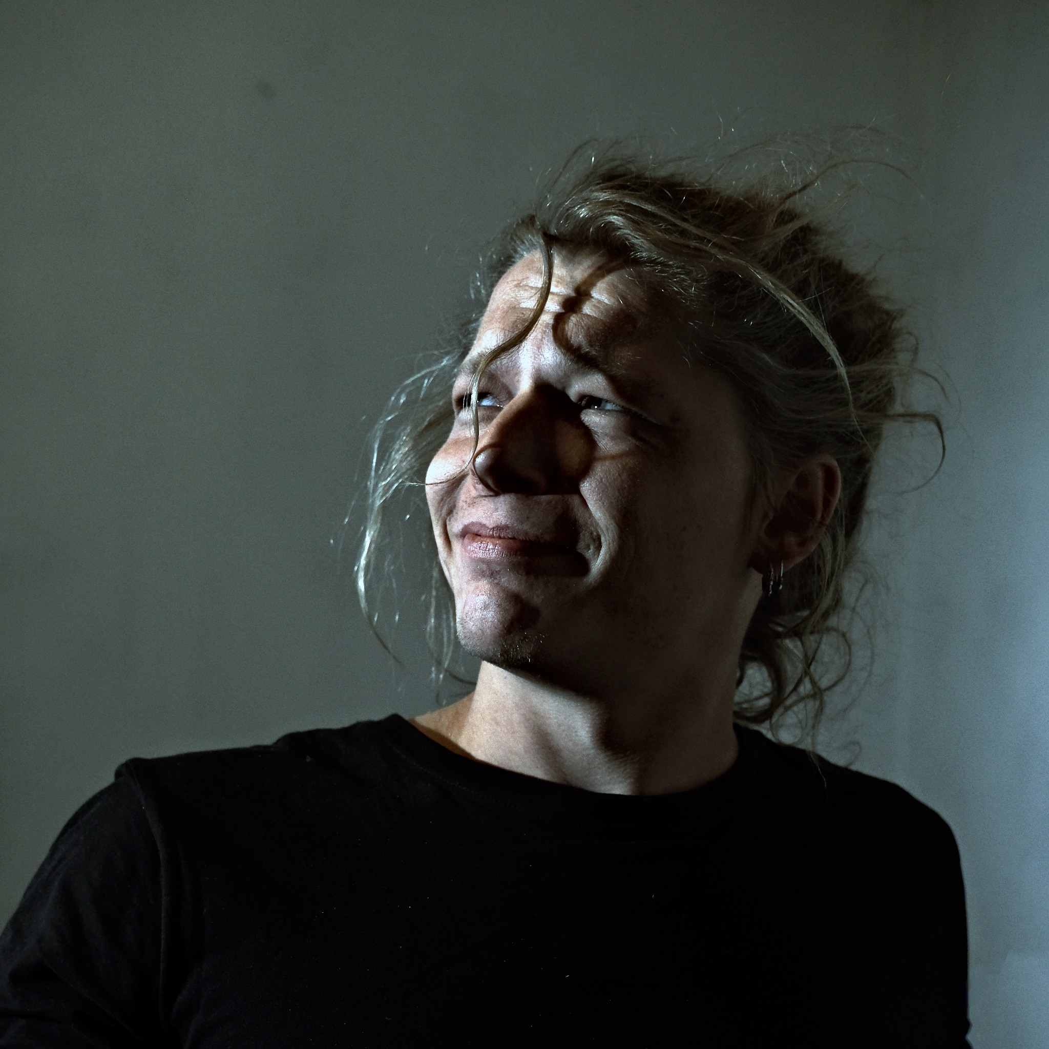 Portret mężczyzny z rozwianymi włosami, zmarszczonym czołem i lekkim uśmiechem, oświetlonego z góry, w czarnej koszulce na tle szarej ściany.