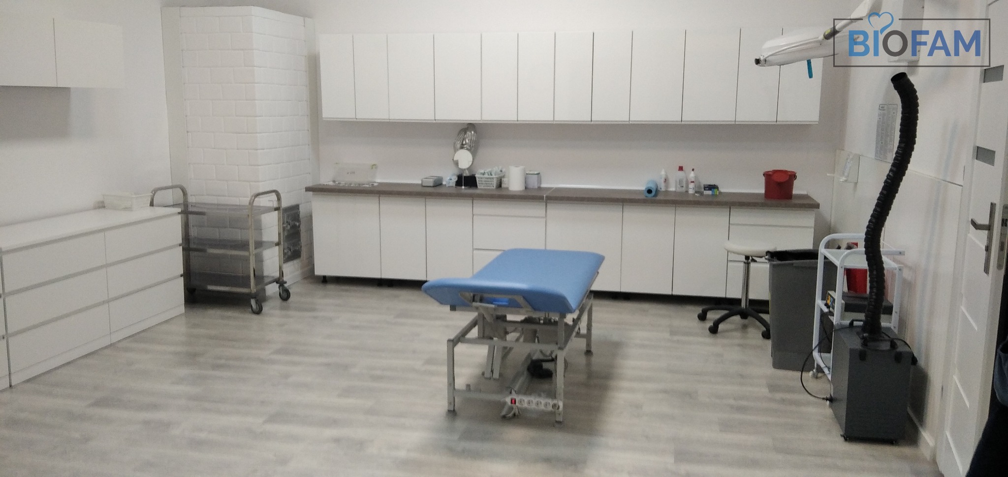 Jasne wnętrze gabinetu medycznego z niebieskim leżanką, białymi szafkami, wózkiem na kółkach i urządzeniem z czarnym wężem ssącym oraz logo Biofam w prawym górnym rogu.