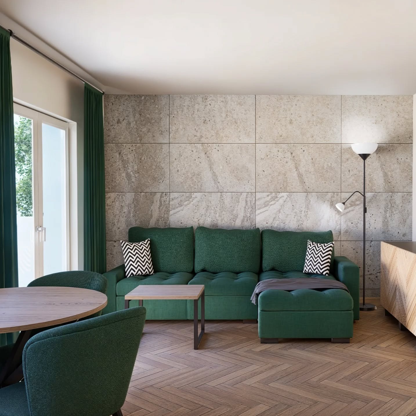 Stylowy salon z zieloną sofą narożną, drewnianym parkietem i ścianą z betonowych płyt. Okno z zielonymi zasłonami i lampa podłogowa dopełniają aranżację.