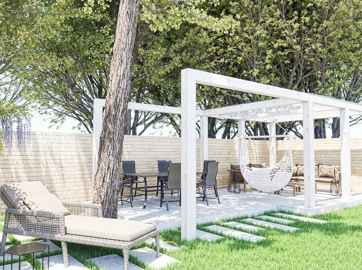 Pergola z huśtawką i meblami ogrodowymi na drewnianym podeście, otoczona zielenią i drewnianym płotem, w słoneczny dzień. Widoczne drzewo wkomponowane w konstrukcję.