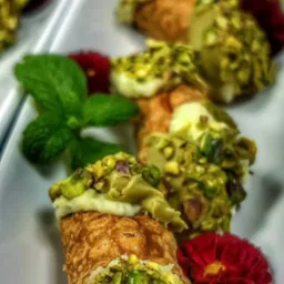 Elegancko podane cannoli z kremem i pistacjami, udekorowane miętą i czerwonym kwiatem na białym talerzu, w tle widoczne kolejne porcje.
