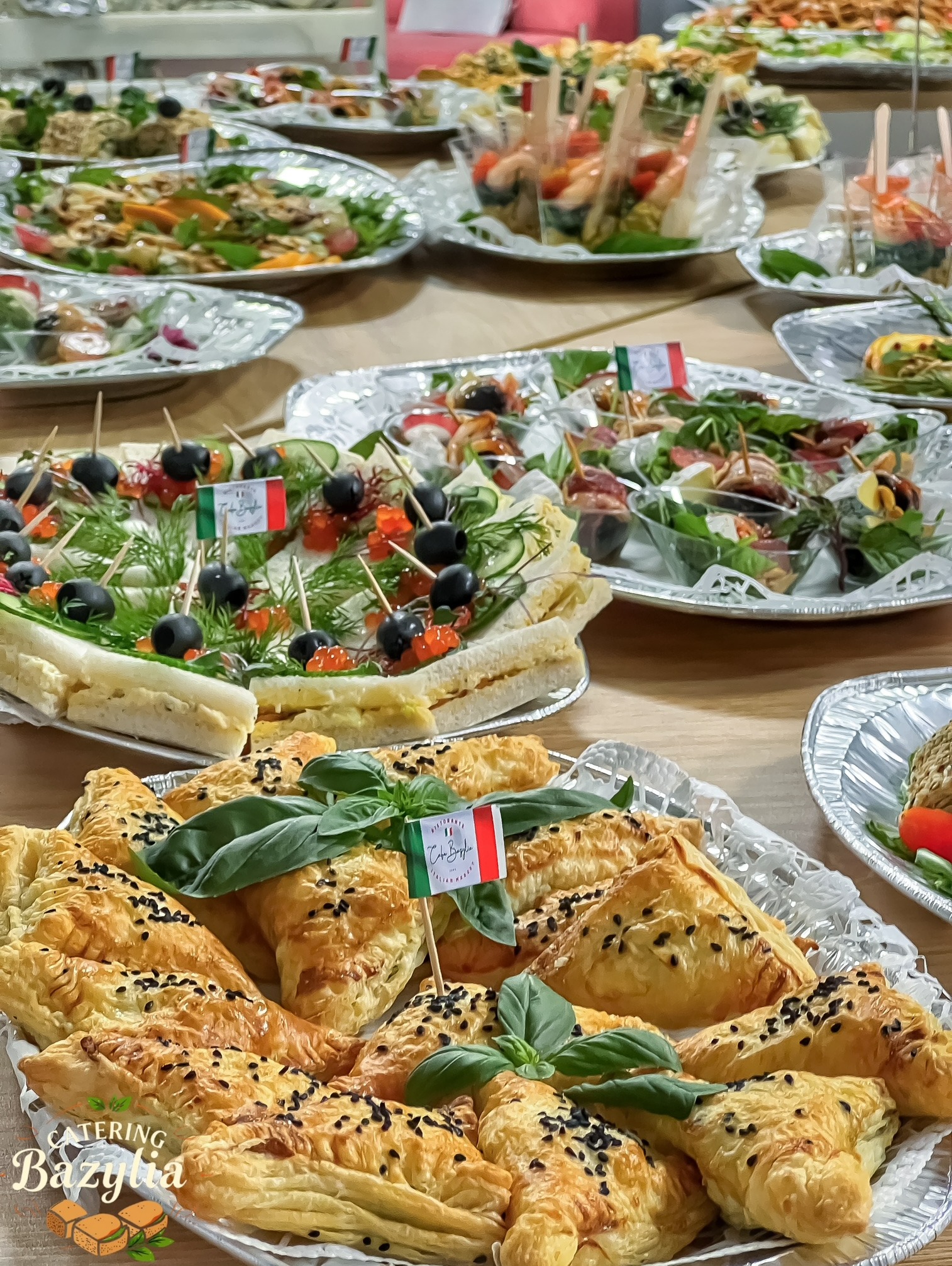 Stół cateringowy z różnorodnymi przekąskami: trójkątne paszteciki z czarnym sezamem i bazylią, kanapki z kawiorem i oliwkami, sałatki z krewetkami, udekorowane flagami Włoch.