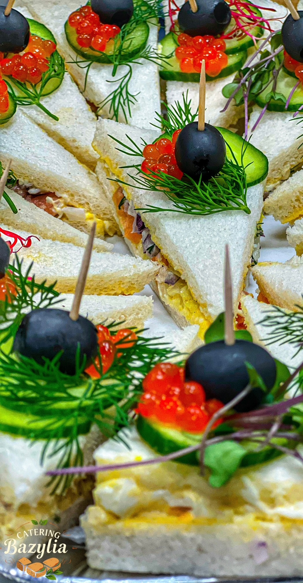 Trójkątne kanapki na białym pieczywie, dekorowane kawiorem, czarną oliwką, ogórkiem i koperkiem, spięte wykałaczkami, ułożone blisko siebie.