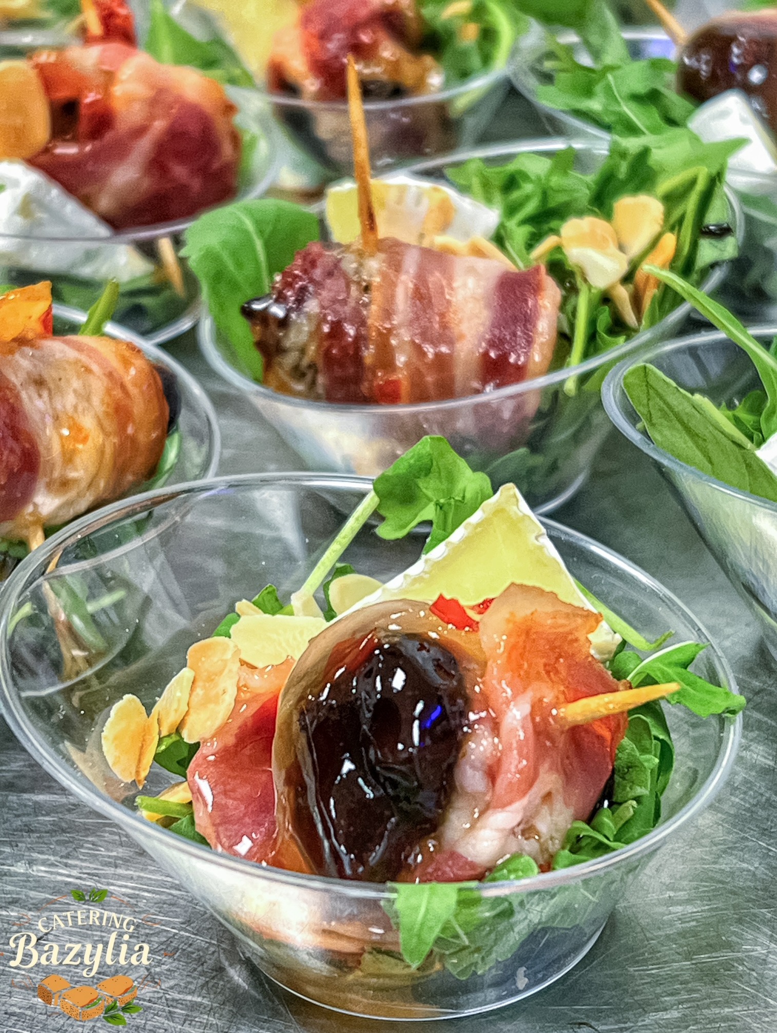 Elegancko podane przekąski w przezroczystych miseczkach: daktyle wędzone w boczku na szpikulcu, ser brie, rukola i płatki migdałów, ułożone w rzędach na szarym tle.