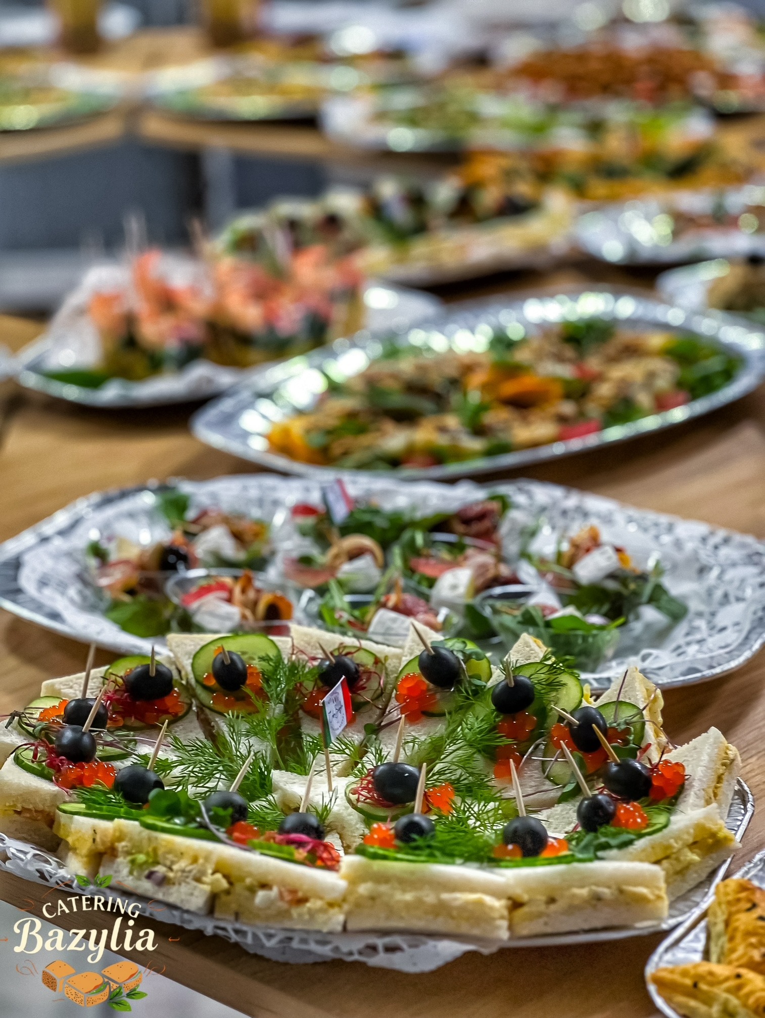 Elegancko podane kanapeczki z kawiorem, oliwkami i ogórkiem na srebrnej tacy, w tle inne dania cateringowe na drewnianym stole.