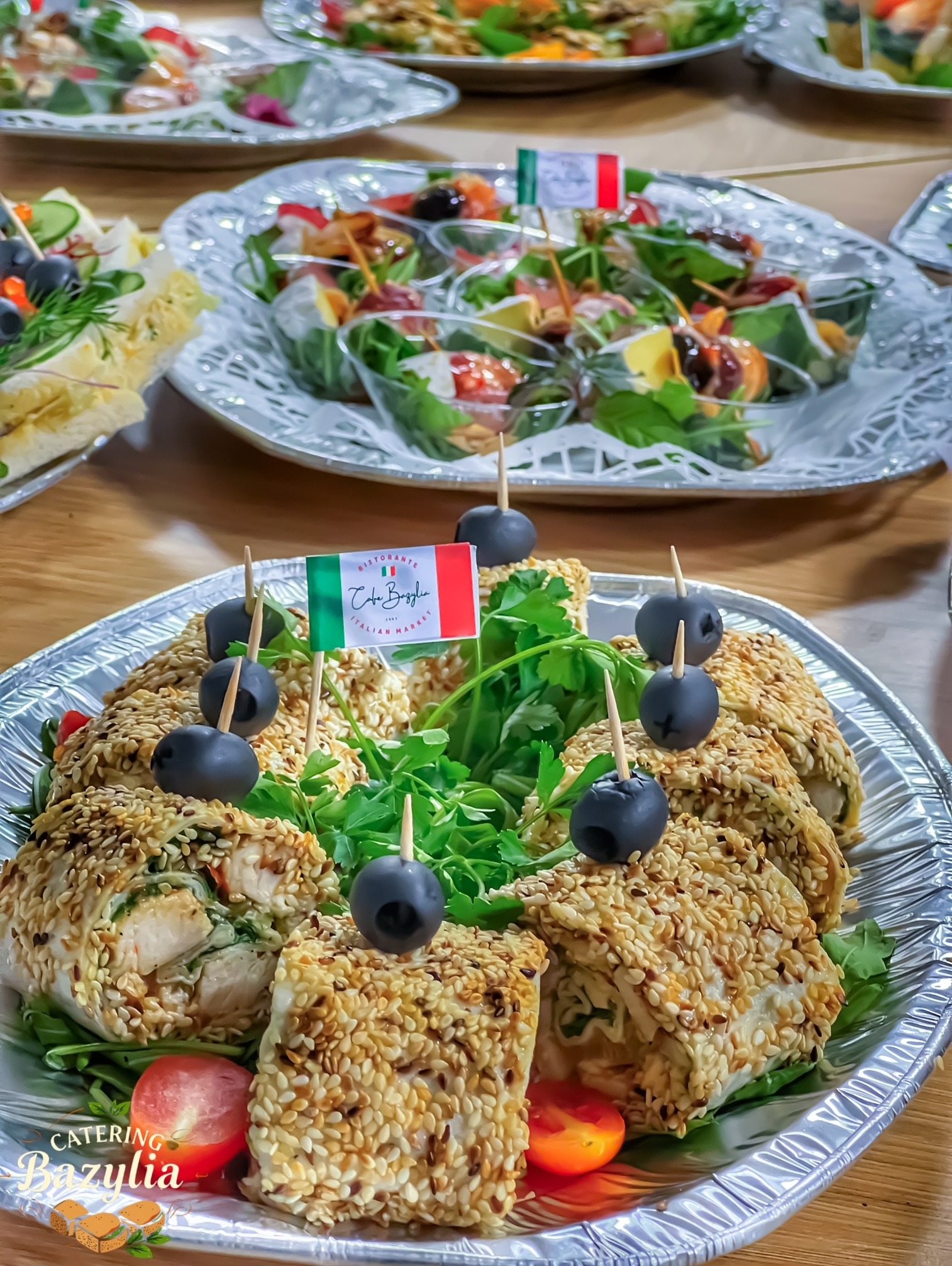 Elegancko podane roladki z kurczaka obtoczone sezamem, udekorowane czarnymi oliwkami na wykałaczkach i świeżą zieleniną, na srebrnej tacy, z małą flagą włoską, w tle inne tace z przekąskami.