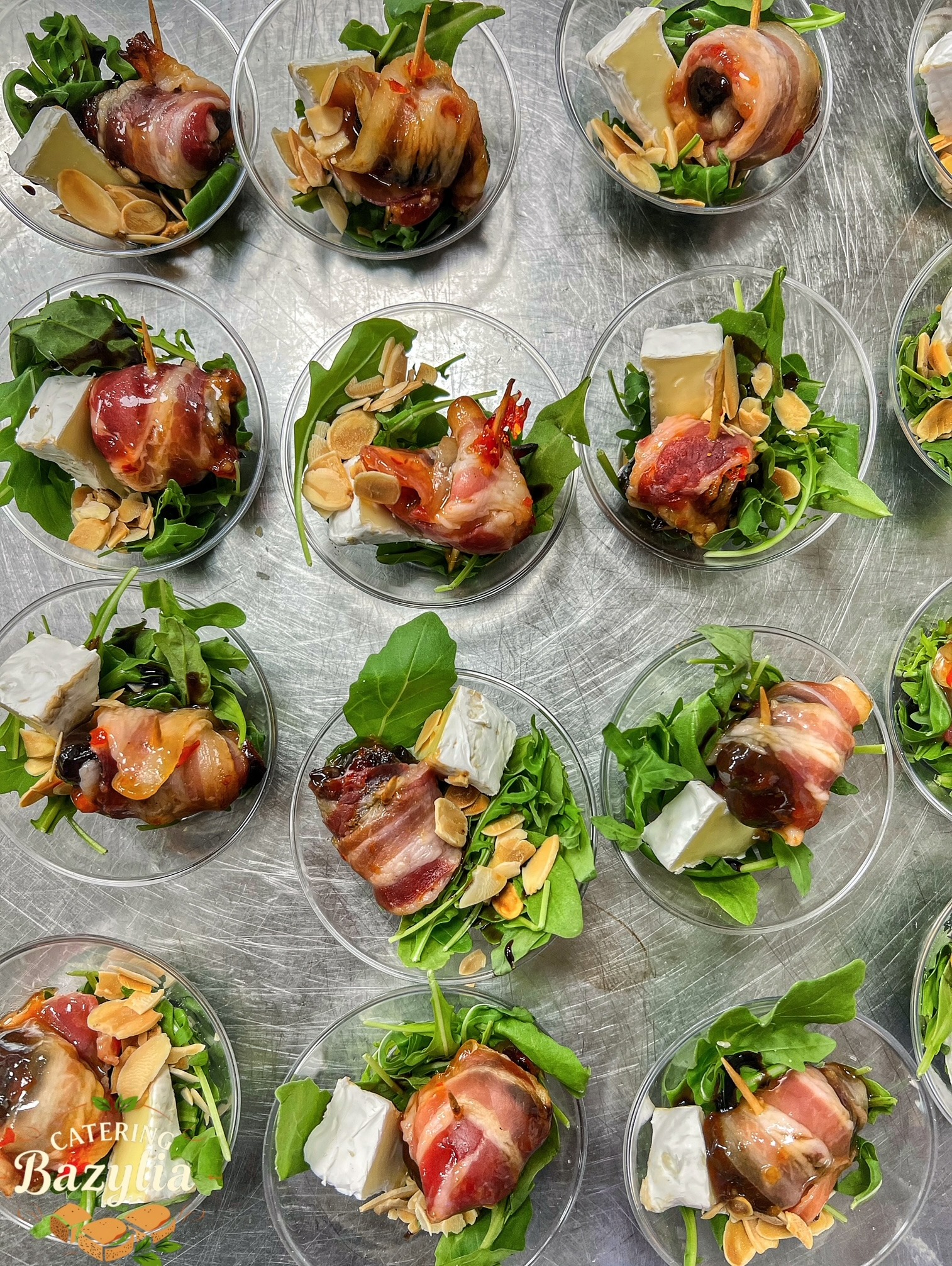 Apetizer w przezroczystym kubeczku: daktyl owinięty boczkiem, ser camembert, rukola i płatki migdałów, całość spięta wykałaczką, na metalowym blacie.