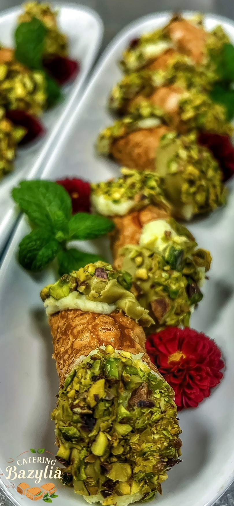 Elegancko podane cannoli z kremem i pistacjami, udekorowane miętą i czerwonym kwiatem na białym talerzu, w tle widoczne kolejne porcje.