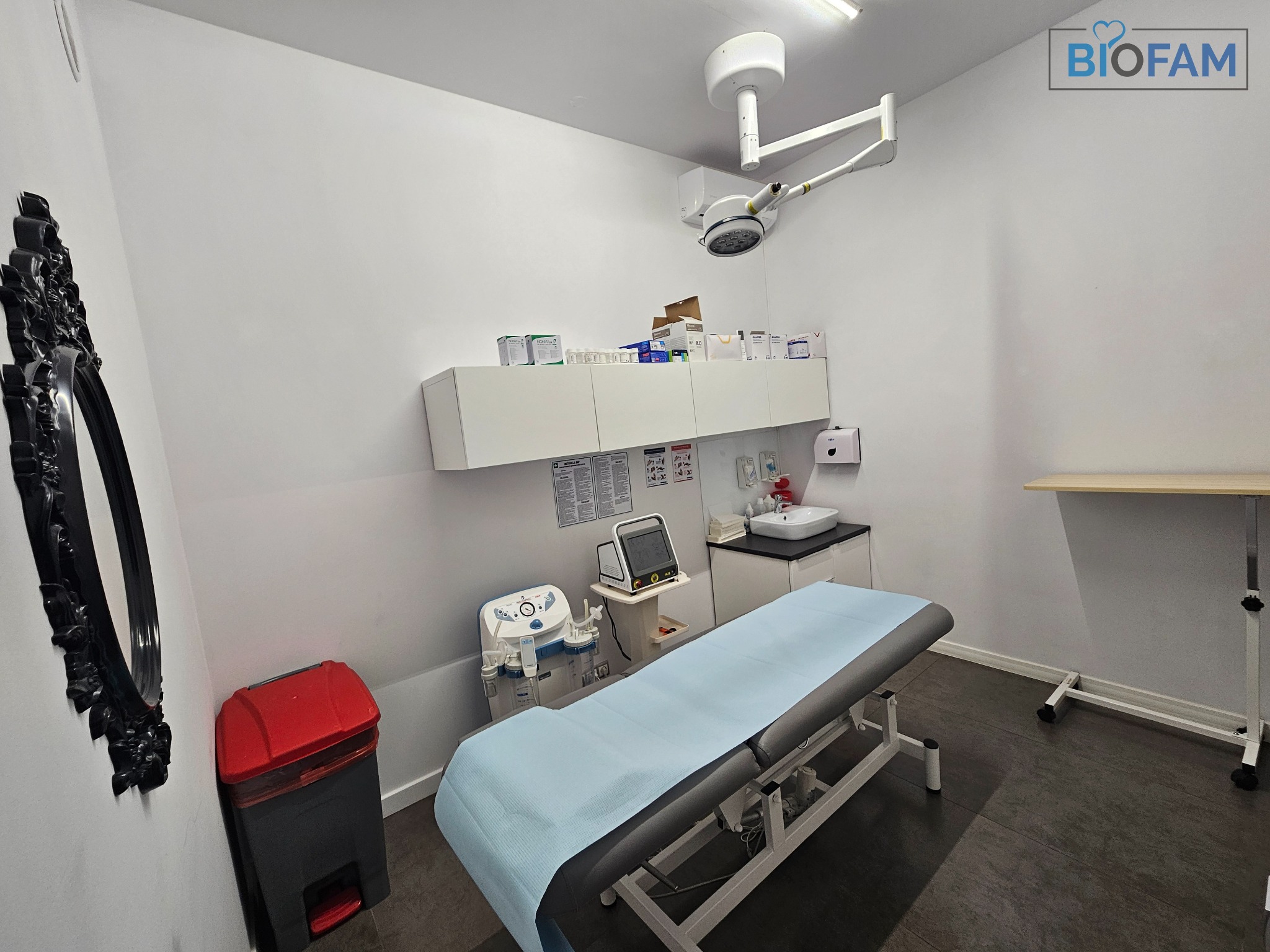 Wnętrze gabinetu lekarskiego z kozetką pokrytą jasnoniebieskim prześcieradłem, sprzętem medycznym, szafkami z lekami, lampą operacyjną i dekoracyjnym lustrem w czarnej ramie.