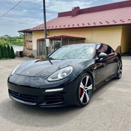 Lasyk Cars - Autodetailing - Czarny samochód marki Porsche Panamera zaparkowany na betonowym placu przed budynkiem z czerwoną, metalową dachówką.