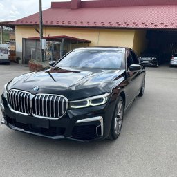 Lasyk Cars - Autodetailing - Czarna limuzyna BMW serii 7, zaparkowana na zewnątrz budynku z czerwonym dachem, z widocznym logo BMW na grillu i odbiciami chmur na lakierze.