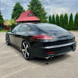 Czarny samochód Porsche Panamera 4S, widok z tyłu, zaparkowany na betonowym placu, w tle altana z czerwonym dachem i rząd zielonych drzew.