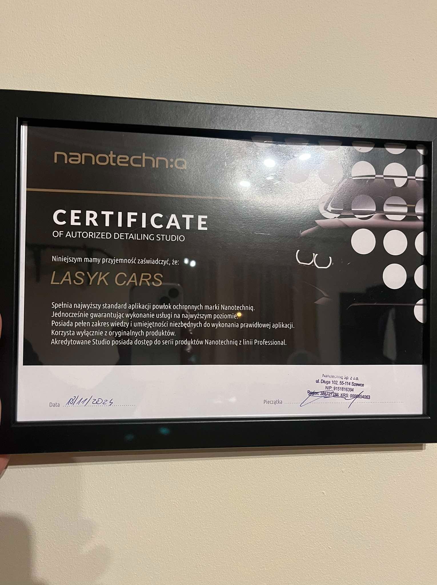 Oprawiony certyfikat autoryzacji studia detailingowego Nanotechniq dla firmy Lasyk Cars, z datą ważności 18.11.2024, wiszący na ścianie.