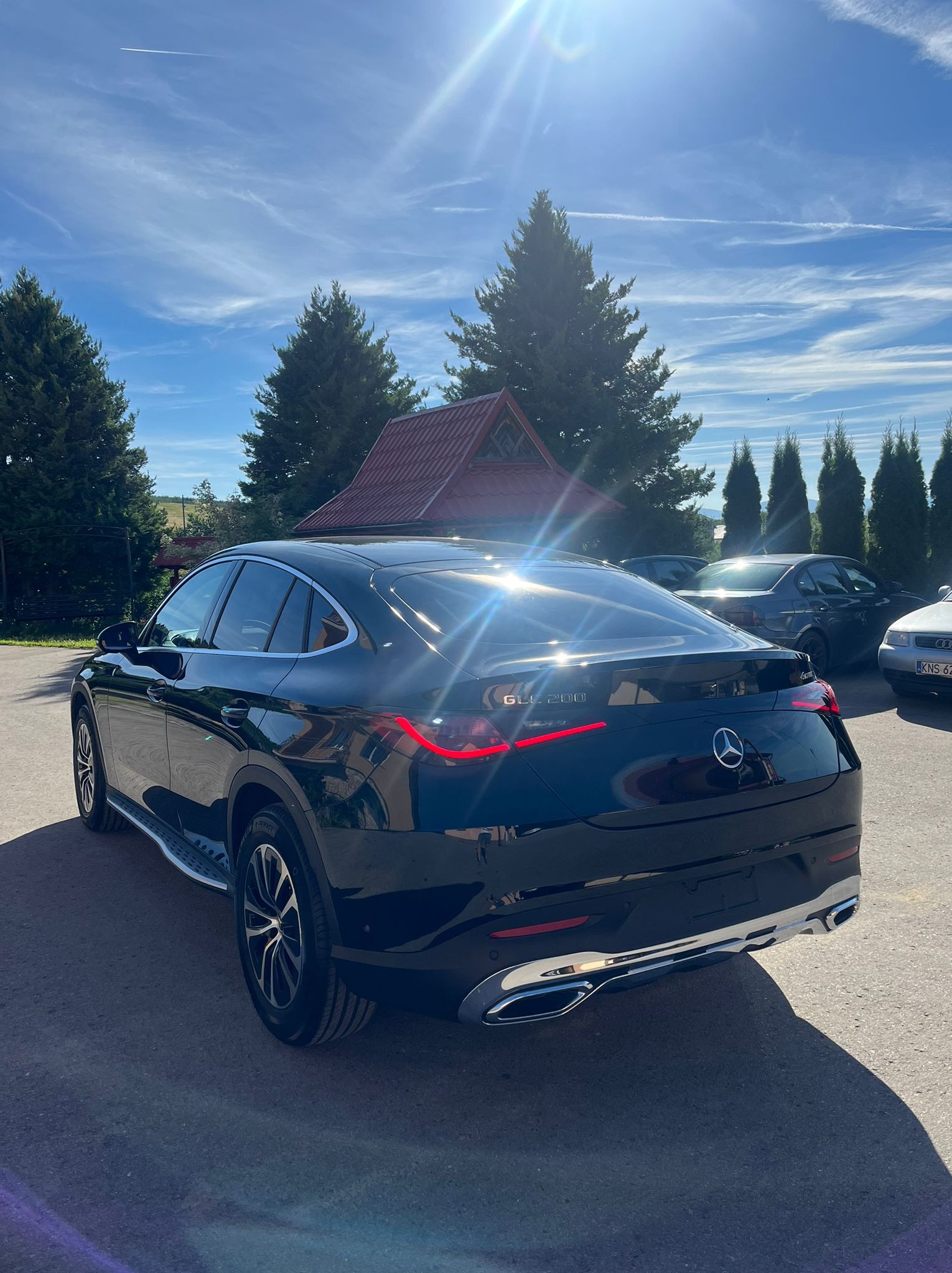 Błyszczący czarny Mercedes-Benz GLC 200 Coupe, widok z tyłu na tle zielonych drzew i błękitnego nieba z chmurami.