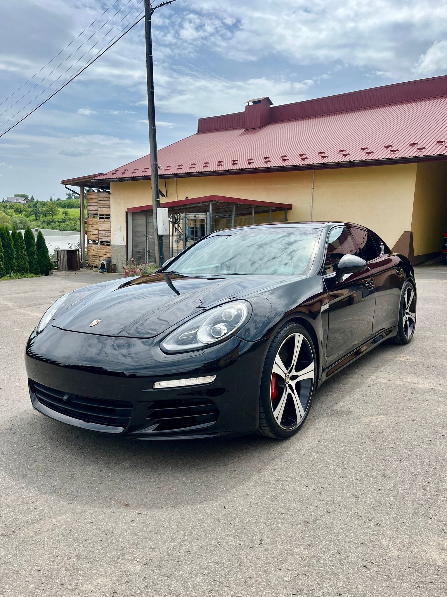 Czarny samochód marki Porsche Panamera zaparkowany na betonowym placu przed budynkiem z czerwoną, metalową dachówką.
