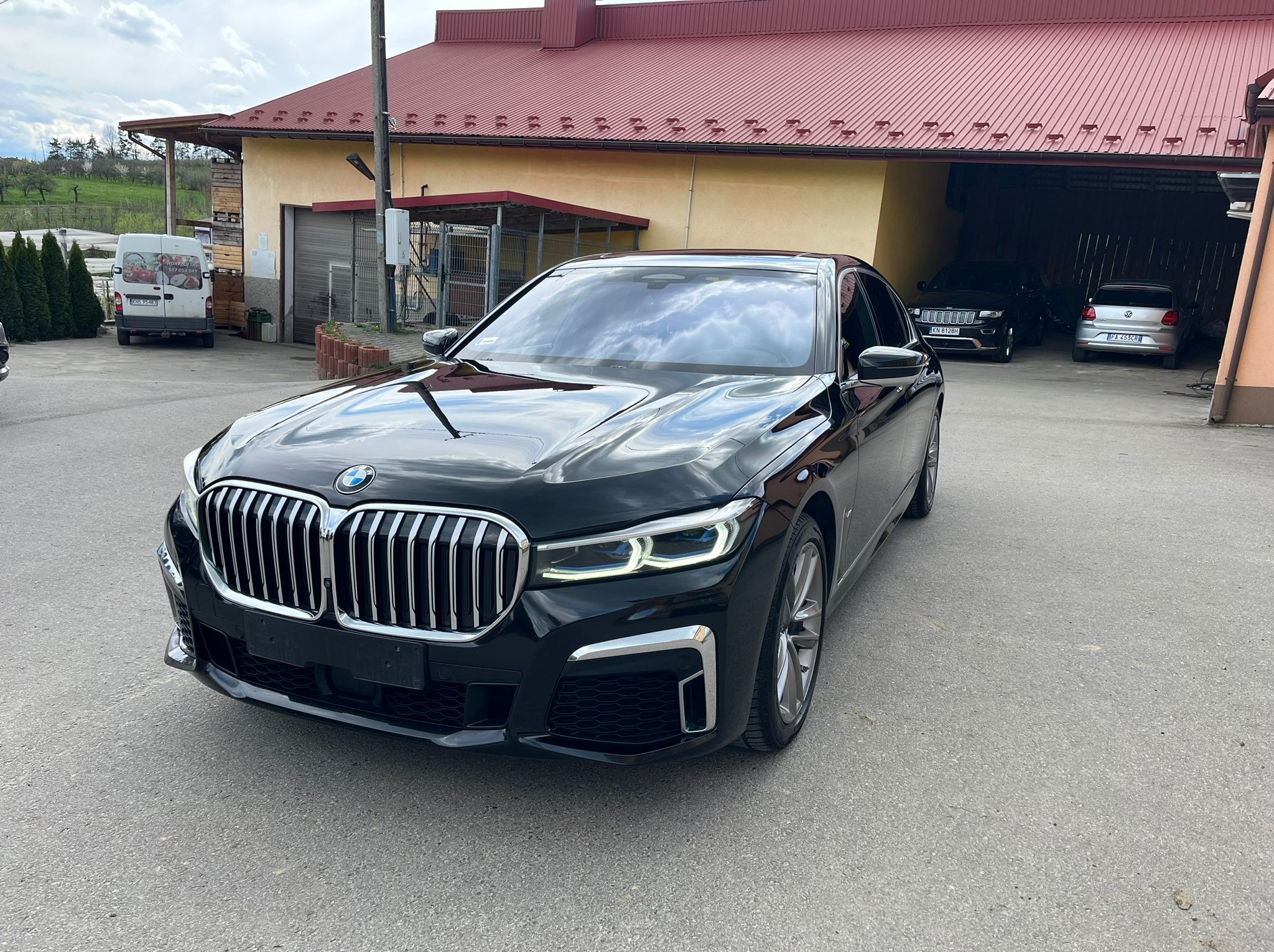 Czarna limuzyna BMW serii 7, zaparkowana na zewnątrz budynku z czerwonym dachem, z widocznym logo BMW na grillu i odbiciami chmur na lakierze.