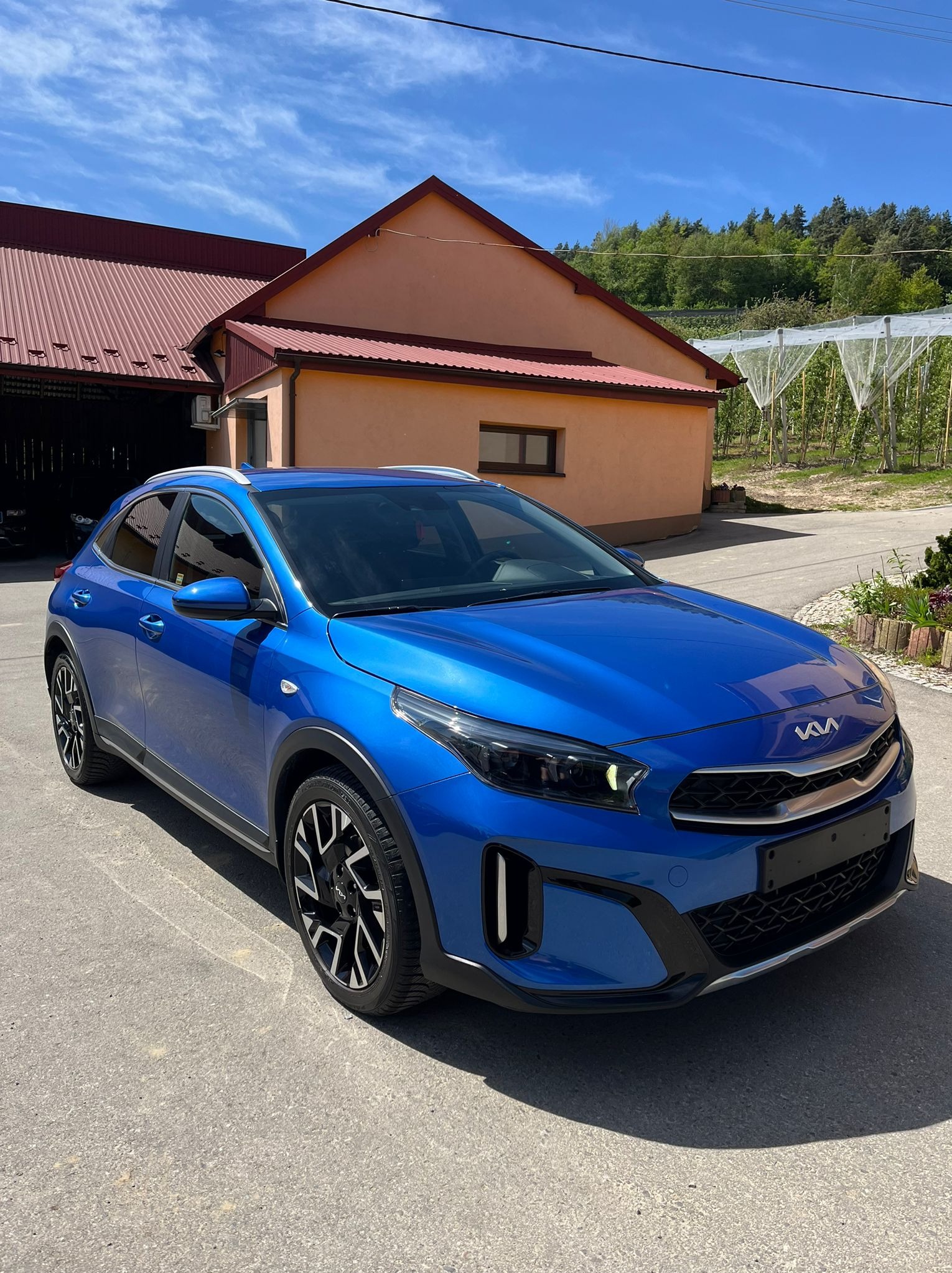 Błękitny samochód Kia XCeed zaparkowany na tle budynku z czerwoną dachówką i sadu z siatkami ochronnymi.