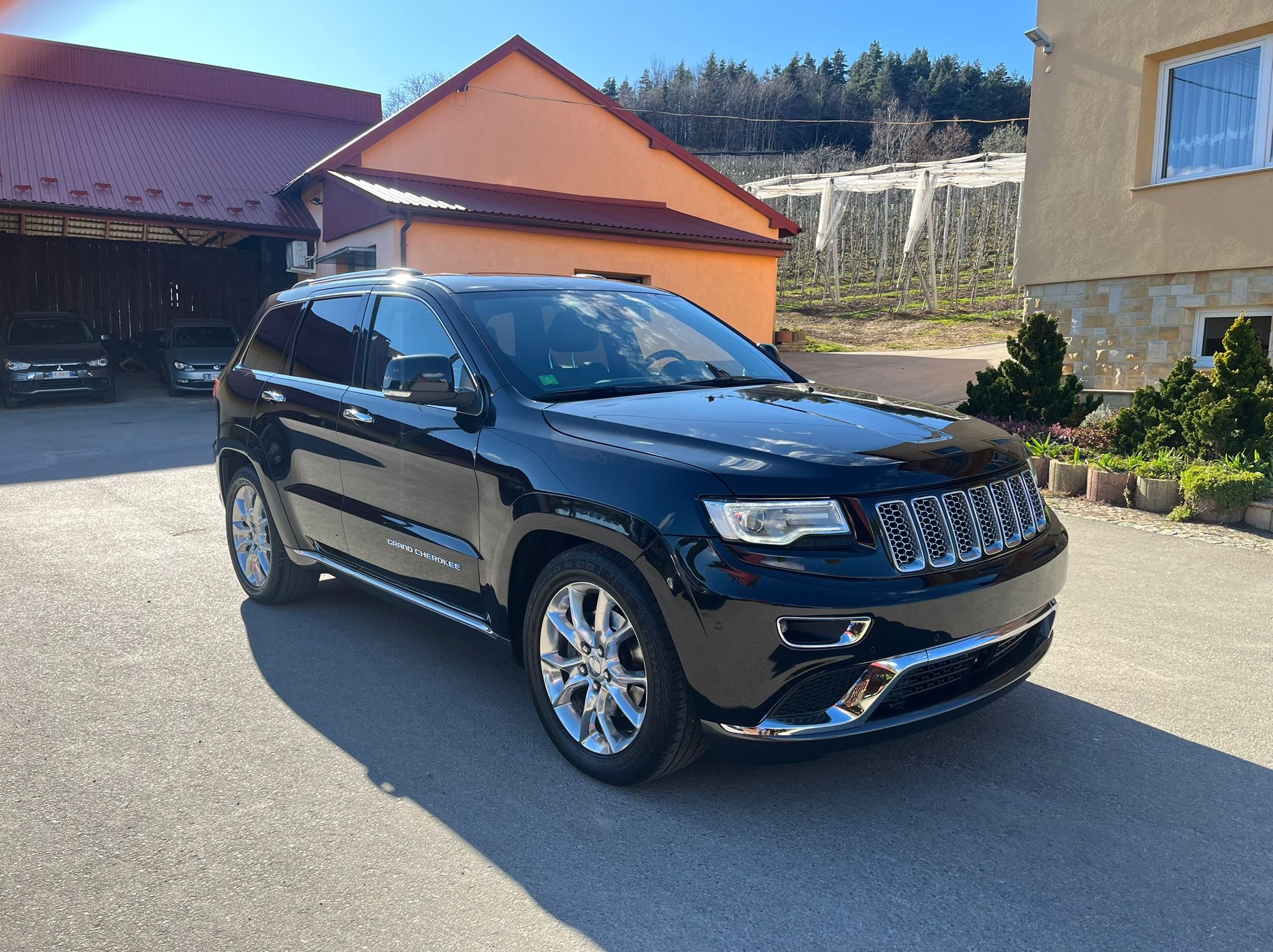 Błyszczący, czarny Jeep Grand Cherokee zaparkowany na asfalcie przed budynkiem z pomarańczową elewacją i czerwoną dachówką w słoneczny dzień.