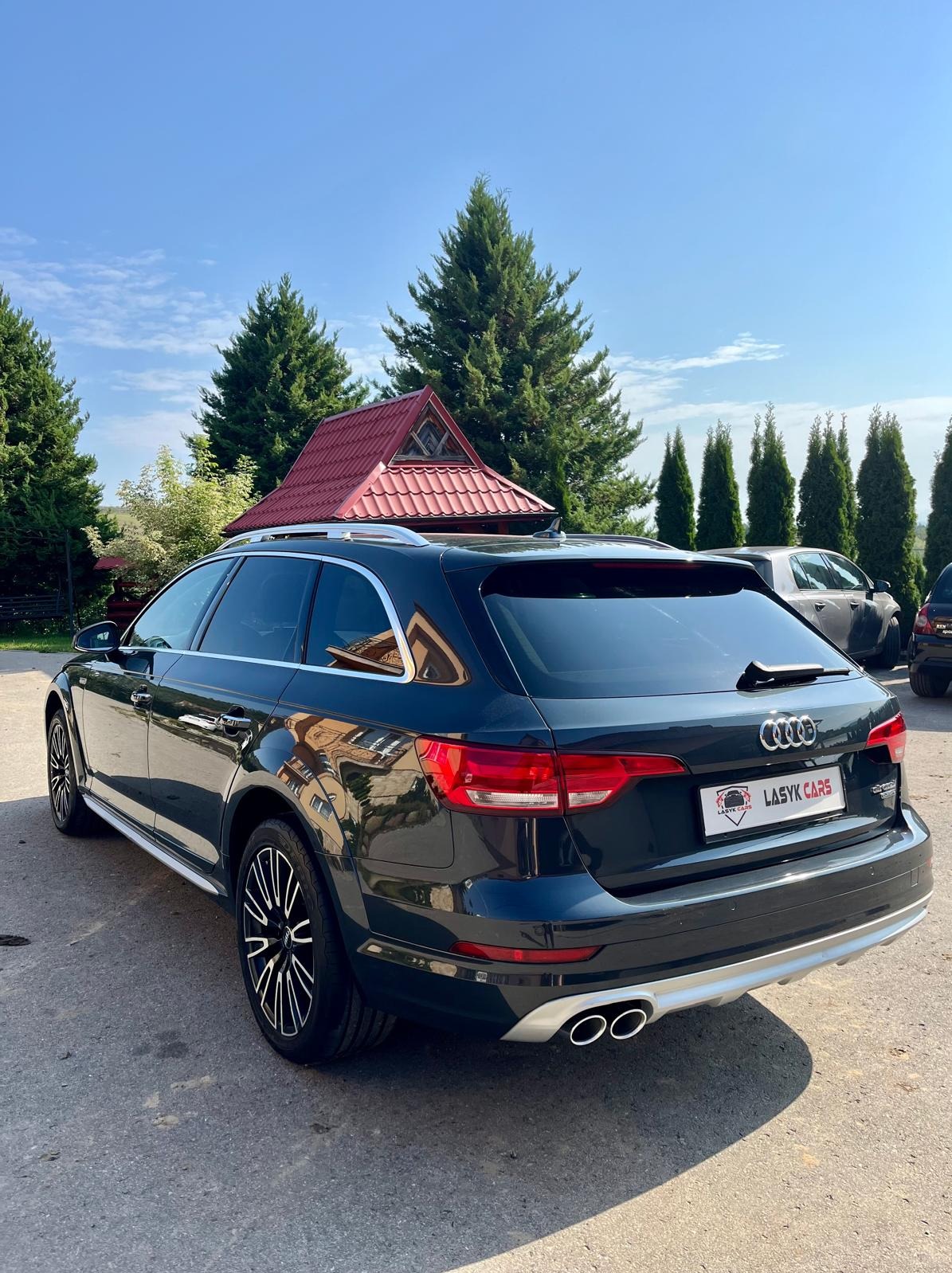 Ciemny samochód Audi A4 Allroad kombi, widok z tyłu, z logo firmy LASYK CARS na tablicy rejestracyjnej, na tle zielonych drzew i czerwonego dachu małej budowli.