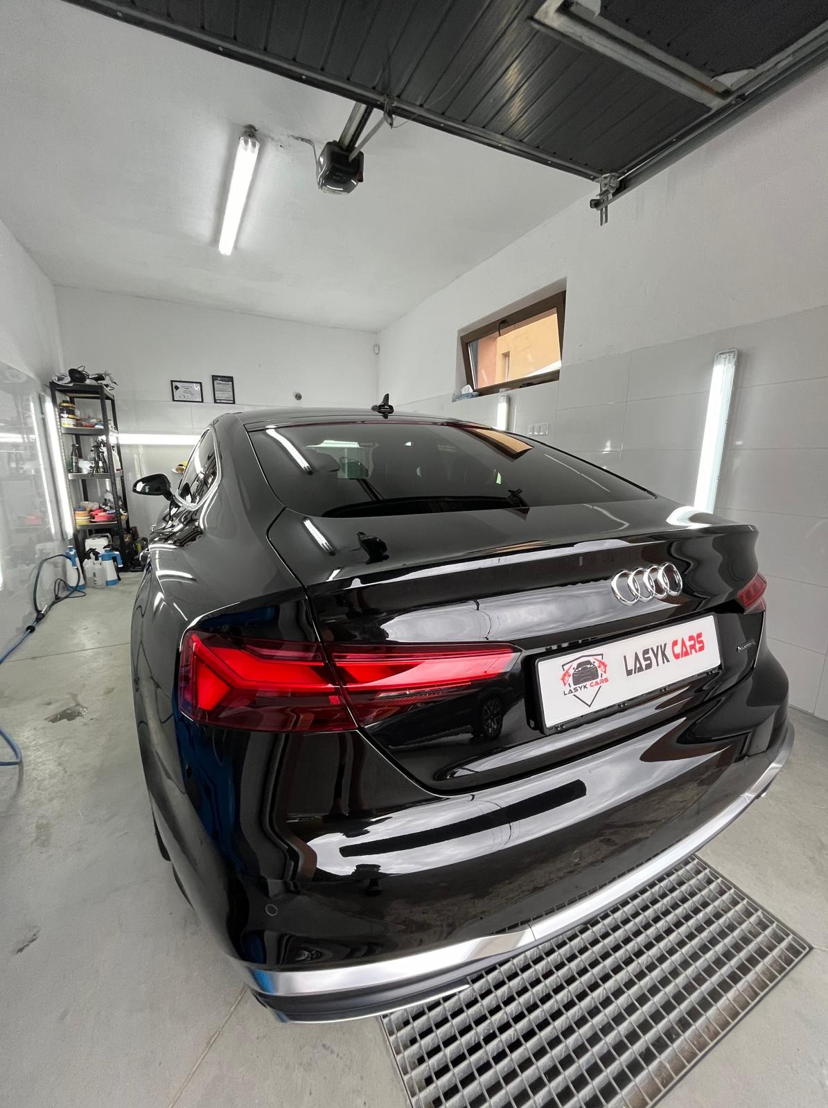 Błyszczący, czarny samochód Audi A5 po umyciu, zaparkowany w dobrze oświetlonym garażu z kratką ściekową pod spodem i półkami z kosmetykami samochodowymi w tle. Widoczna tablica rejestracyjna...