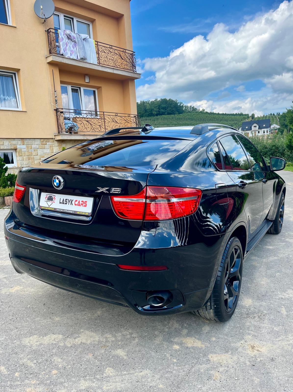 Czarny, lśniący BMW X6 zaparkowany przed domem z beżową elewacją. Na tylnej klapie widoczna tabliczka z logo firmy LASYK CARS.
