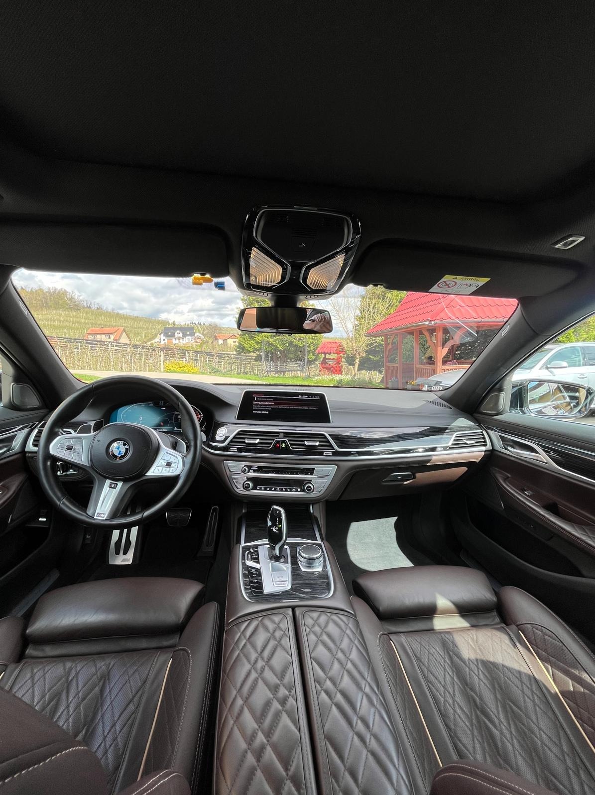 Wnętrze luksusowego samochodu z brązową skórzaną tapicerką, widok na deskę rozdzielczą, kierownicę BMW i fragment widoku na zewnątrz z czerwoną altaną w tle.