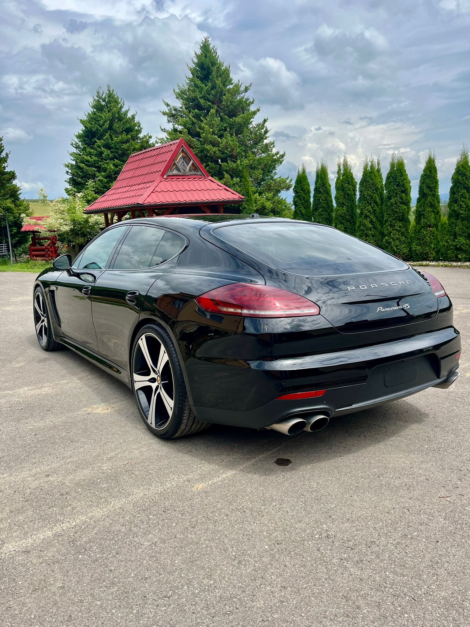 Czarny samochód Porsche Panamera 4S, widok z tyłu, zaparkowany na betonowym placu, w tle altana z czerwonym dachem i rząd zielonych drzew.