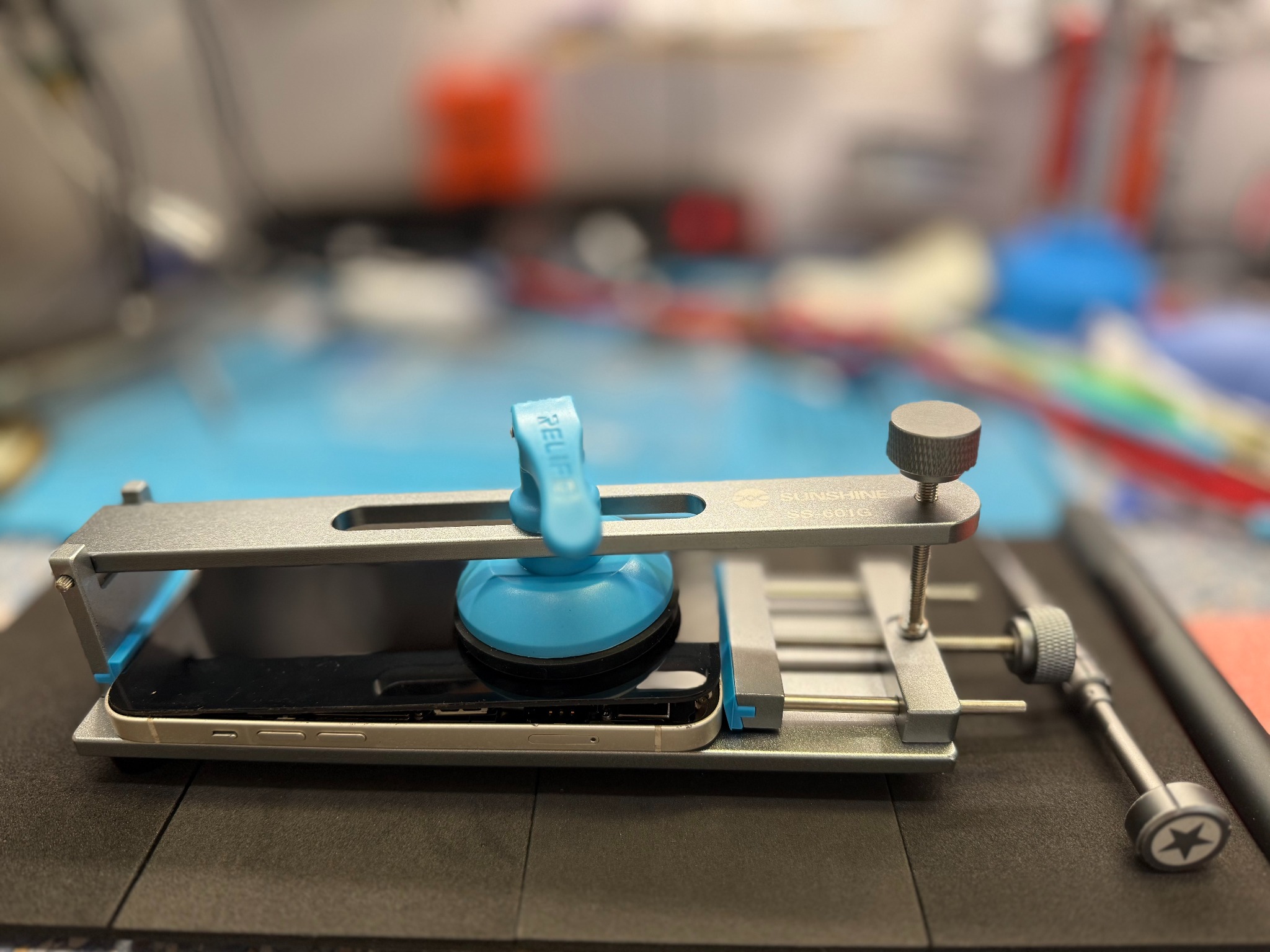 Telefon zamocowany w metalowej ramie z przyssawką podczas demontażu, widoczne narzędzia na stole warsztatowym.