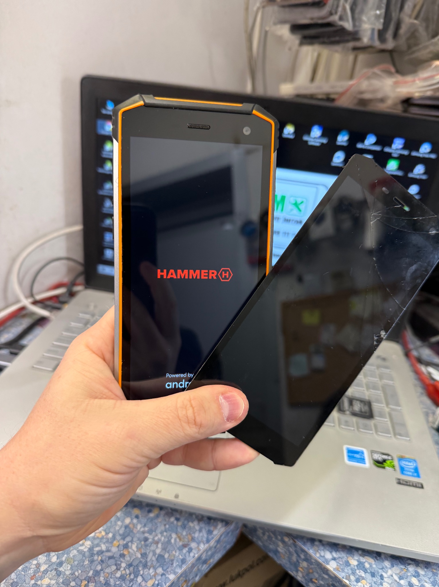 Dłoń trzyma telefon marki Hammer z włączonym ekranem startowym obok uszkodzonego ekranu innego telefonu na tle otwartego laptopa.