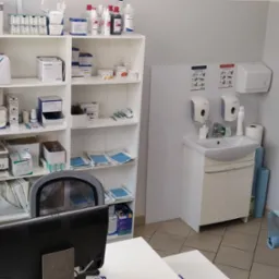 Jasne wnętrze gabinetu lekarskiego z umywalką, szafką z licznymi lekami i materiałami medycznymi na półkach oraz kozetką z niebieskim podkładem.