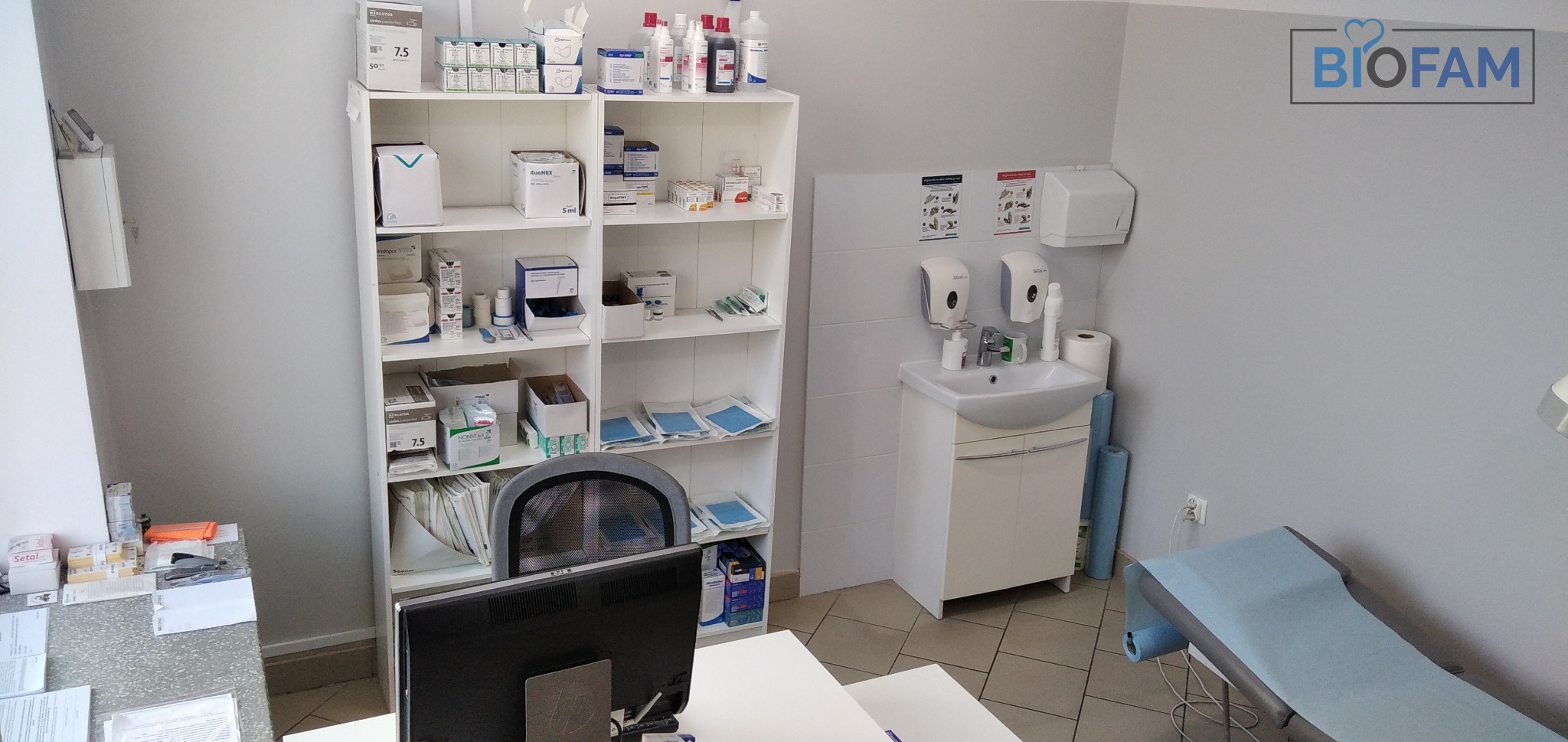 Jasne wnętrze gabinetu lekarskiego z umywalką, szafką z licznymi lekami i materiałami medycznymi na półkach oraz kozetką z niebieskim podkładem.