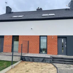 Nowoczesny dom jednorodzinny z elewacją w kolorach bieli, szarości i czerwonej cegły. Dach z oknami połaciowymi, antena satelitarna i komin.