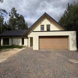 AIA.MAKS.GROUP - Nowoczesny dom jednorodzinny z garażem w Grodzisku Mazowieckim. Jasna elewacja, grafitowy dach, drewniana brama garażowa. Widok z przodu, żwirowe podłoże.