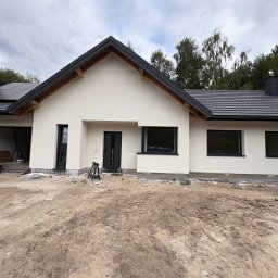 AIA.MAKS.GROUP - Nowoczesny dom z białą elewacją, czarną stolarką okienną i dachem z panelami solarnymi. Widoczne ślady prac wykończeniowych na posesji.