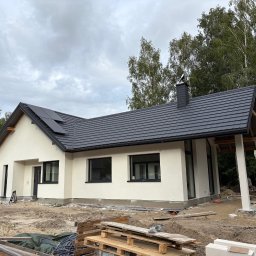 AIA.MAKS.GROUP - Nowoczesny dom jednorodzinny z jasną elewacją i ciemną dachówką, częściowo ocieplony, z widocznymi panelami słonecznymi na dachu i elementami wykończeniowymi.