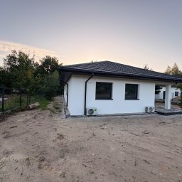 AIA.MAKS.GROUP - Biały dom z czarnym dachem i oknami, dwa klimatyzatory na elewacji, widok z zewnątrz, teren wokół domu w trakcie zagospodarowywania, ogrodzenie w tle.
