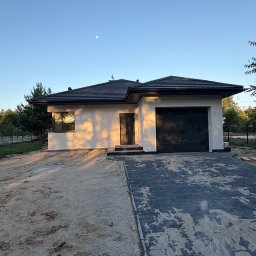 AIA.MAKS.GROUP - Nowoczesny dom jednorodzinny z garażem, wykończony jasnym tynkiem, w promieniach zachodzącego słońca. Widoczny fragment podjazdu z kostki brukowej i ogród.