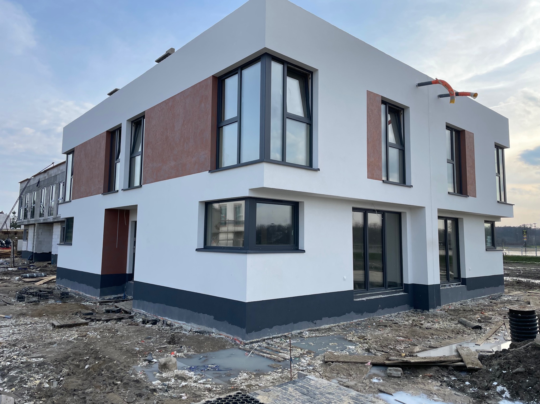 Nowoczesny, dwukondygnacyjny dom w trakcie wykańczania elewacji, z białymi ścianami, brązowym akcentem i szarym cokołem, widoczne instalacje na zewnątrz, teren wokół w stanie budowy.
