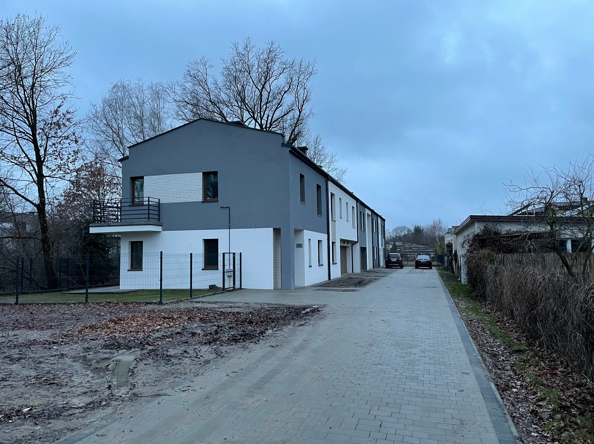 Nowoczesny szeregowiec z szarą i białą elewacją, widok z poziomu ulicy w pochmurny dzień. Widoczne detale wykończenia, takie jak balustrada balkonu i numer domu '23D'.