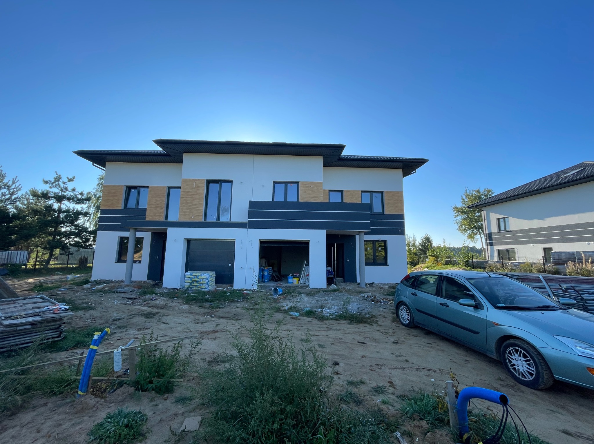 Dwukondygnacyjny dom bliźniak w trakcie wykańczania elewacji, widoczne worki z materiałami budowlanymi, niebieski samochód osobowy i elementy instalacji na trawniku przed budynkiem.