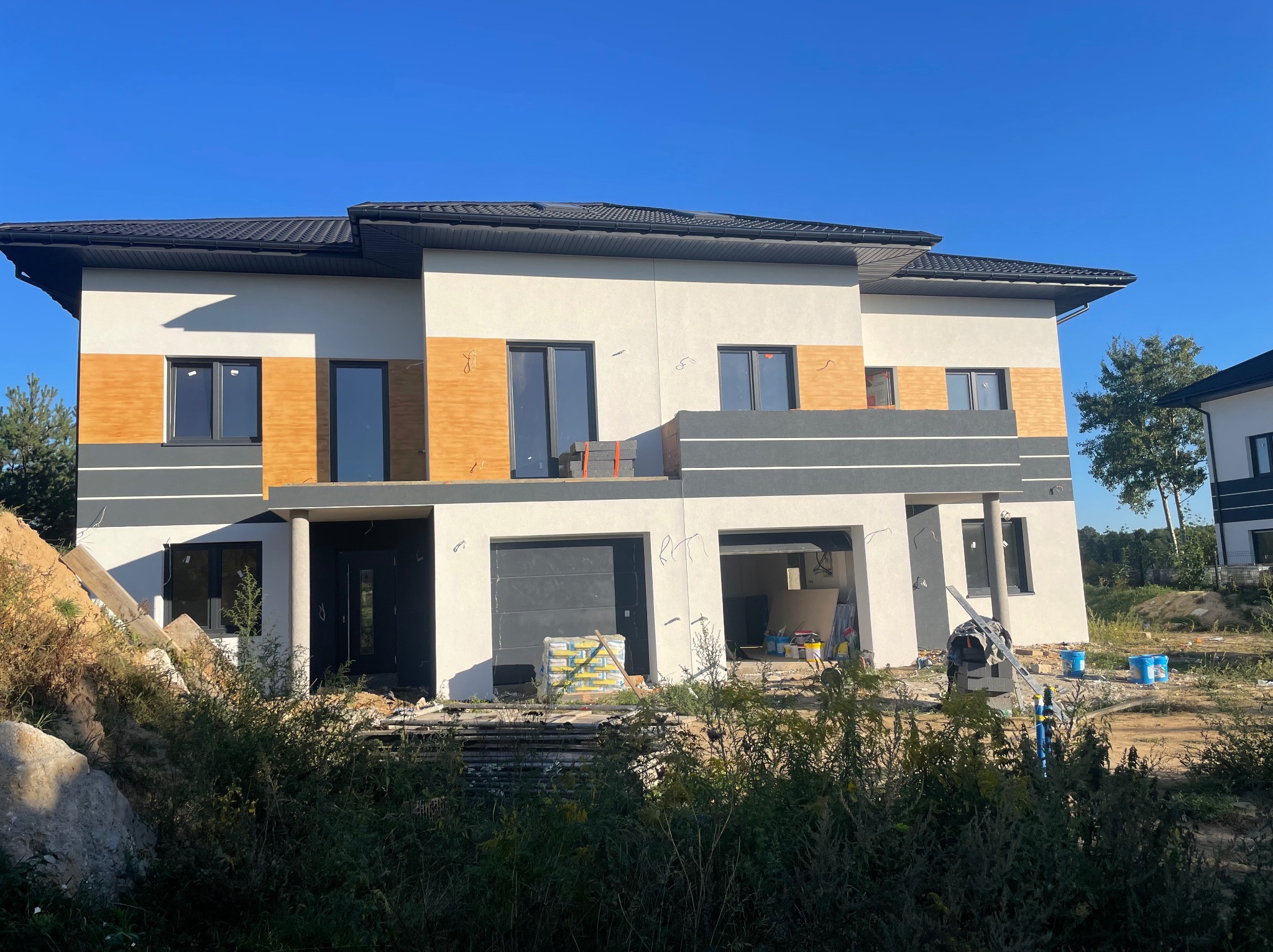 Dwukondygnacyjny dom w trakcie wykonywania elewacji, z widocznymi fragmentami ocieplenia i tynku, na tle błękitnego nieba i zarośli.