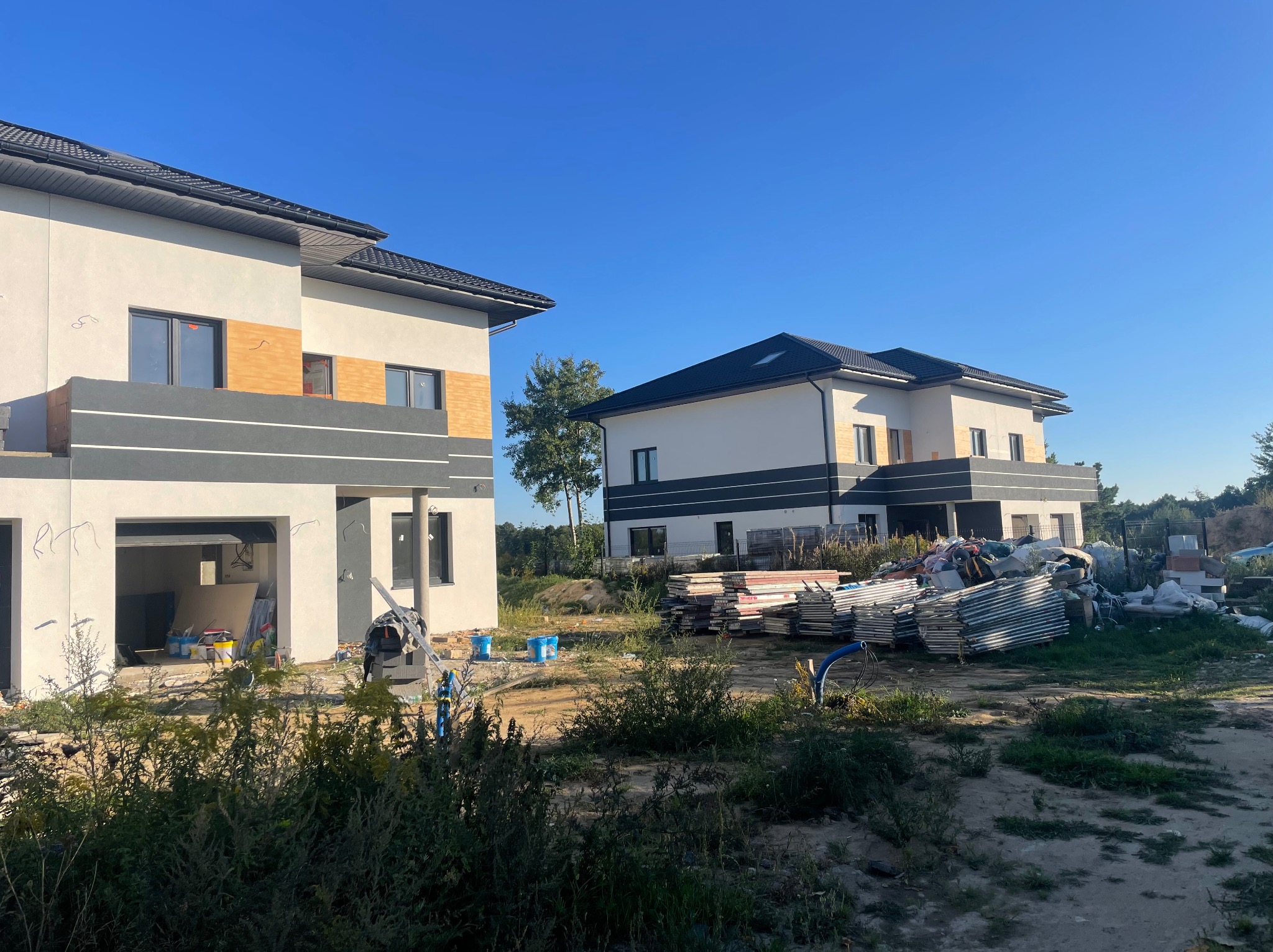 Dwa nowoczesne domy w stanie surowym zamkniętym, z częściowo wykonaną elewacją w kolorze białym i szarym, widoczne materiały budowlane i narzędzia na działce, błękitne niebo w tle.