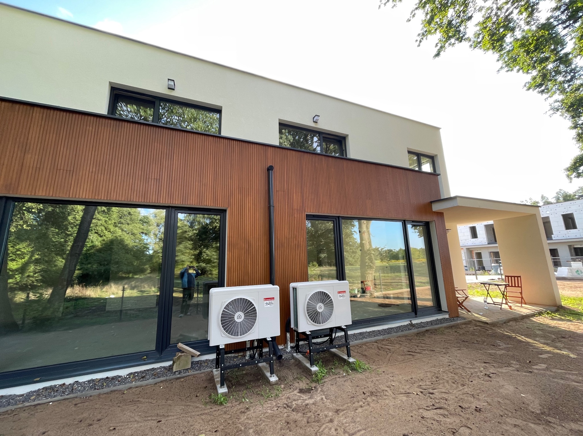 Nowoczesny dom z elewacją w kolorze kremowym i brązowym, z dużymi oknami i zainstalowanymi dwoma zewnętrznymi jednostkami klimatyzacji na metalowych podstawach przed budynkiem.