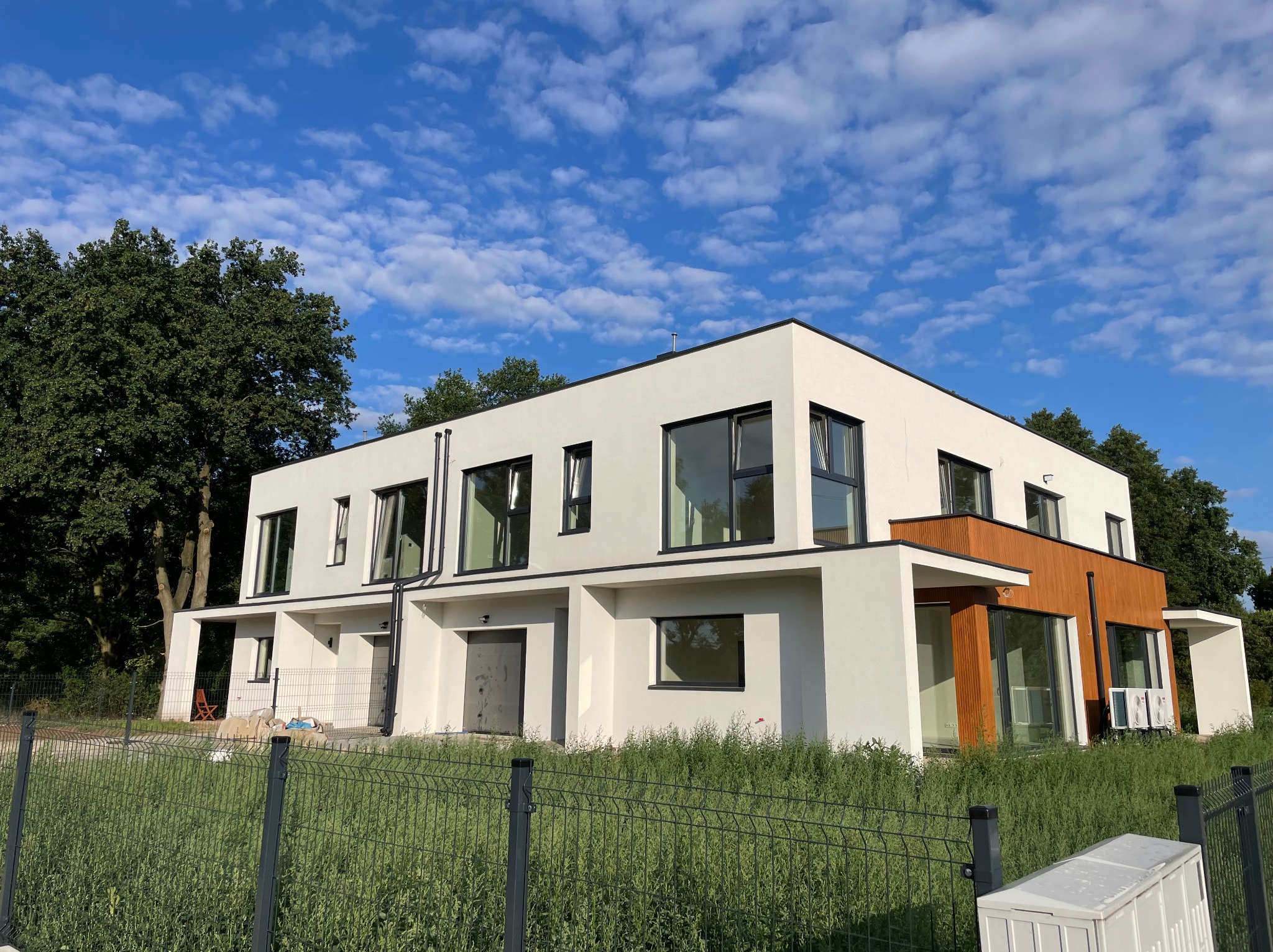 Nowoczesny dwukondygnacyjny dom z białą elewacją i drewnianymi panelami, widoczny zza ogrodzenia zarośniętego wysoką trawą, na tle błękitnego nieba z obłokami.