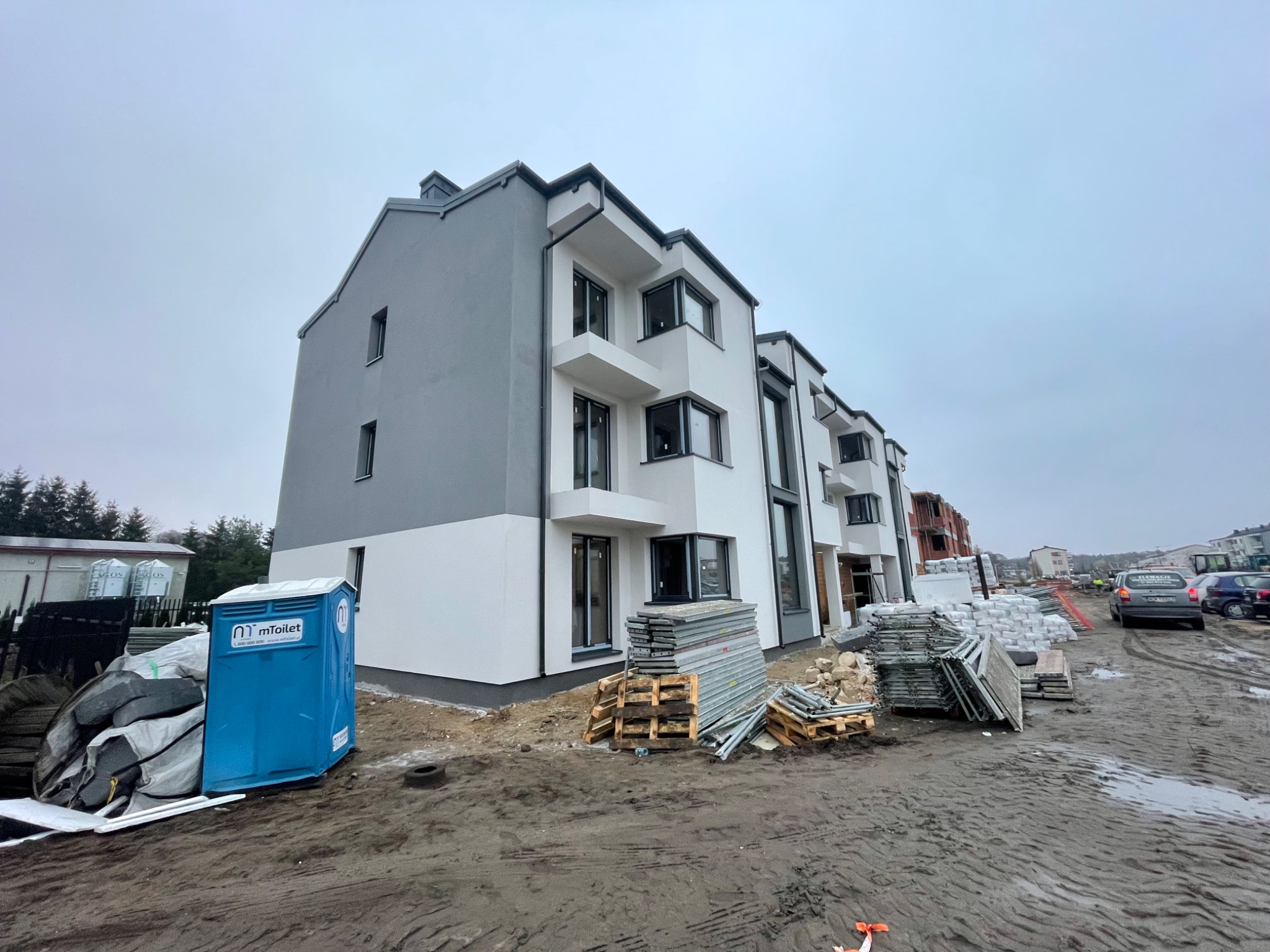 Nowoczesny budynek wielorodzinny z szarą i białą elewacją, z widocznymi rusztowaniami i przenośną toaletą. Ujęcie z dołu podkreśla bryłę budynku.